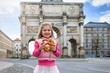 A cute girl holding a traditional, german pretzel in front of the Victory Gate in Munich, Germany, as a ravel concept