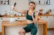 Pretty young woman with slender body using modern smartphone for filming video about healthy eating. Happy blonde in stylish sport outfit sitting on chair and holding glass with fresh fruit cocktail.
