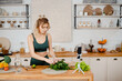 Charming strong woman in green sport outfit recording video on mobile about healthy food. Fitness blogger with blond hair standing on kitchen and making tutorial.