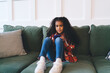 © BullRun - Young African American girl, approximately 6-8 years old, sitting on a green sofa holding a smartphone. She appears focused and calm, wearing a red flannel, blue jeans, and white sneakers