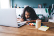Serious African American curly haired female child studying and taking notes in notebook while sitting at wooden table with laptop against blurred background in living room
