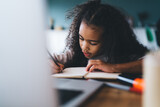 Fototapeta Nowy Jork - Serious African American curly haired female child studying and taking notes in notebook while sitting at wooden table with laptop against blurred background in living room