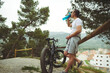 © Taras Grebinets - Caucasian young man cyclist in white t-shirt and sportswear, drinking water from sports bottle, standing by a wooden fence, relaxing after riding an eco-friendly electric bike