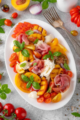 Wall Mural - Caprese salad with mozzarella, tomatoes, prosciutto and grilled peaches on a white plate on a gray concrete background. Fresh summer salad. Top view.