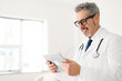 A senior doctor with a gentle smile reviews data on a tablet, a sign of embracing modern medical technology in patient care, side view. Healthcare and technology concept