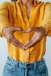 A close-up of a person's hands creating a heart shape, dressed in a vivid yellow shirt, symbolizing love and acceptance.