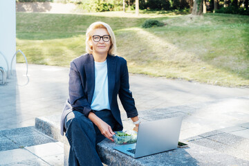 Wall Mural - Business woman eating fast and healthy in front of laptop during job lunch break outdoors. Modern business lifestyle. Middle aged lady eats salad and use notebook pc on bench. Balanced diet lunch box.