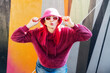 © okrasiuk - Hipster young woman with pink hair and sunglasses in magenta fluffy sweatshirt and bucket hat posing on the wall background. Urban street fashion. Mono color look. Gen Z, millennials self expression.
