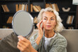 © Юлия Завалишина - Love yourself. Beautiful old mature woman enjoying mirror reflection. Senior adult older lady looking at mirror confident happy on sofa at home. Woman doing daily morning beauty routine. Self love