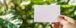 Closeup hand holding blank white card mockup on blurred green plant background, interior design with copy space area for text