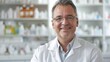 A man in a white lab coat is smiling for the camera. He is standing in front of a pharmacy with shelves full of medicine