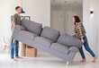 Happy young couple moving sofa in living room, replacing furniture at home, copy space. Millennial husband and wife making rearrangement in their apartment, side view