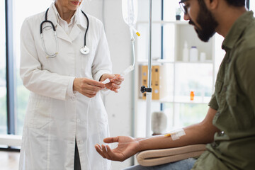 Wall Mural - Efficient general practitioner checking solution for chemotherapy of male sitting on exam couch. Young bearded man in casual wear taking dropper while mature female making system review.