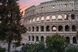 Fototapeta  - Ancient amphitheater in Rome, Italy, showcasing iconic Colosseum exterior with arches, stone walls, and greenery. Tourists and locals walk near historic site at twilight, capturing serene vibe.