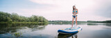 Fototapeta  - A young woman with an open swimsuit swims on a SUP board on a picturesque lake. Evening tour. A beautiful, slender girl is engaged in sap surfing on the calm water of a picturesque pond.