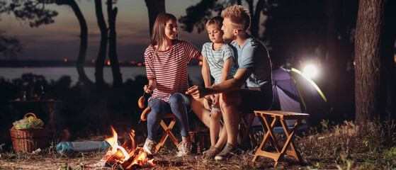 Wall Mural - Family roasting sausages over campfire in evening