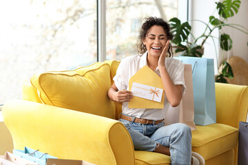 Canvas Print - Happy young African-American woman with gift card in envelope sitting on sofa at home