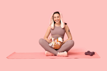 Wall Mural - Happy female fitness coach in bunny ears with Easter basket and dumbbells on pink background