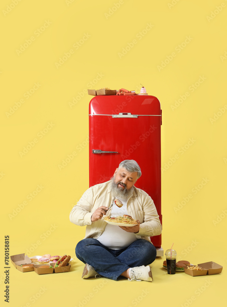 Overweight mature man with unhealthy food sitting near fridge on yellow background. Overeating concept