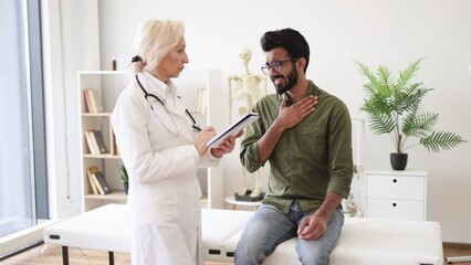 Wall Mural - Caucasian senior female doctor and young male patient sitting at exam couch of modern medical center and diagnose disease. Elder specialist writing instruction for taking medication correctly.