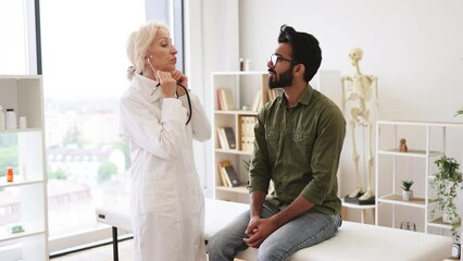 Wall Mural - Side view of focused elderly lady in white coat using stethoscope while young man sitting on exam couch. General practitioner examining heartbeat while providing regular checkup in clinic.