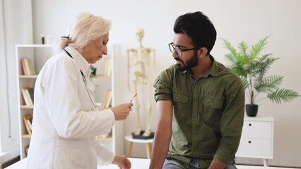 Wall Mural - General practitioner giving injection to boost immunity for young bearded patient. Confident mature woman in lab coat holding syringe with seasonal flu vaccine while young man sitting in exam room.