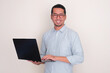 © KrishnaTedjo - Adult Asian man smiling at the camera while holding a laptop