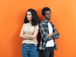 © puhhha - Conflict And Resolution: Young Multiethnic Couple Standing Back To Back, Looking Upset Against An Orange Background. Representing Relationship Issues, Diversity, And Emotion.