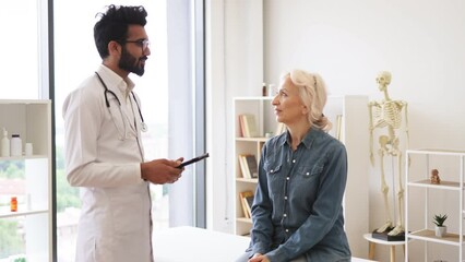 Wall Mural - Portrait of bearded physician using tablet for writing electronic prescription. Senior woman describing symptoms of illness to young male doctor at modern medical office, looking, at camera.