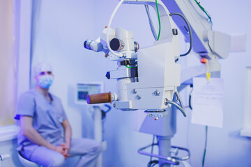 An oculist doctor sits on the background of a modern microscope for vision testing in the office. Medical room for vision treatment