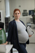 Pregnant woman standing and holding documents on a paper tablet in the office. Vertical photo. 