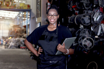 Wall Mural - Portrait of African American woman with curly hair wears apron, holds laptop computer, stands at auto spare parts store warehouse, surrounded by secondhand engine parts. Female staff at workplace.