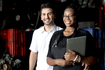 Wall Mural - Portrait of happy harmony people working together at workplace, smiling African American woman holds laptop with her colleague at auto spare parts store warehouse, surrounded by secondhand engine part