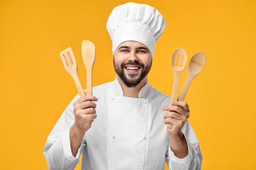 Wall Mural - Happy young chef in uniform holding wooden utensils on orange background