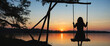 Silhouette of a girl sitting on a swing over the lake at sunset.