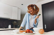Happy African American woman enjoying music while holding a cucumber in the kitchen, smiling and feeling joyful