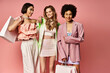 Three women of diverse backgrounds stand side by side, holding shopping bags against a vibrant pink studio backdrop.
