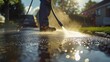 Man power washing a driveway creating a spray of water droplets with a car parked in the background and a house in the distance under a clear blue sky.