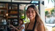 © Z A C - Smiling woman with long hair wearing apron standing in front of a coffee shop with a blurred background of a sunny day.