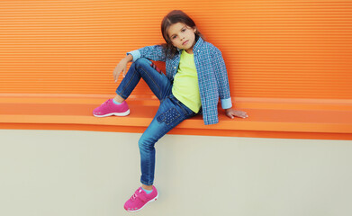 Wall Mural - Stylish girl child in colorful clothes posing on city street on orange wall background