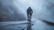 A lone cyclist clad in a black jacket and helmet pedaling down a foggy mountain road surrounded by a rocky cliff and a gray overcast sky.