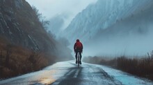 A lone cyclist in a red jacket pedaling down a misty mountain road with steep cliffs on either side.