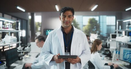 Wall Mural - Portrait of an Indian Adult Male Scientist Using Tablet Computer, Looking at Camera and Smiling in a Laboratory. Professional Researcher Working as Bioengineer, Monitoring Technology Projects