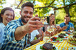 A group of friends having dinner together at an outdoor table, smiling and taking selfies while eating food with red wine on the side