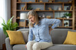 © Liubomir - Mature lady feeling spinal muscles pain attack and holding back while squinting eyes on sofa with cushions. Sick woman having problem with standing up and experiencing kidney ache in living room.