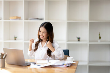 Cheerful businesswoman celebrates a successful moment at her workplace, with a laptop and paperwork on the desk, expressing joy and satisfaction.