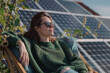 © Femmes.Digital - A relaxed woman in a green sweater and sunglasses lounging on a wooden chair near photovoltaic panels, enjoying the sun at her home rooftop garden with a solar system installation. Earth Day theme.