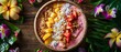 A top view of a tropical smoothie bowl topped with sliced fruits, granola, and coconut flakes, surrounded by tropical foliage and colorful flowers, on a wooden table