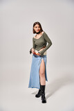Fototapeta  - A young woman with brunette hair poses stylishly in a skirt and boots in a studio setting.