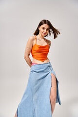 Canvas Print - A young woman with brunette hair poses gracefully in a studio, wearing an orange top and a flowing blue skirt.